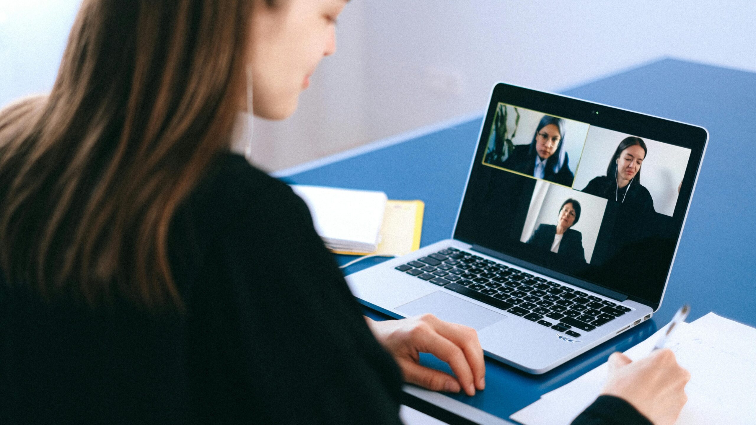 A woman engaging in a video conference using a laptop at home, taking notes.
